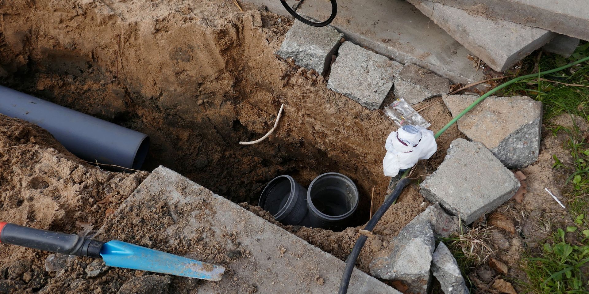 A trench in sand with gray pipes, paving stones, and a garden tool.