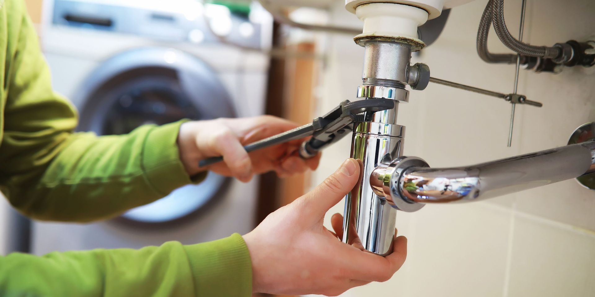 Person in green shirt using a wrench to fix a sink's plumbing in a bathroom. A washing machine is visible in the background.