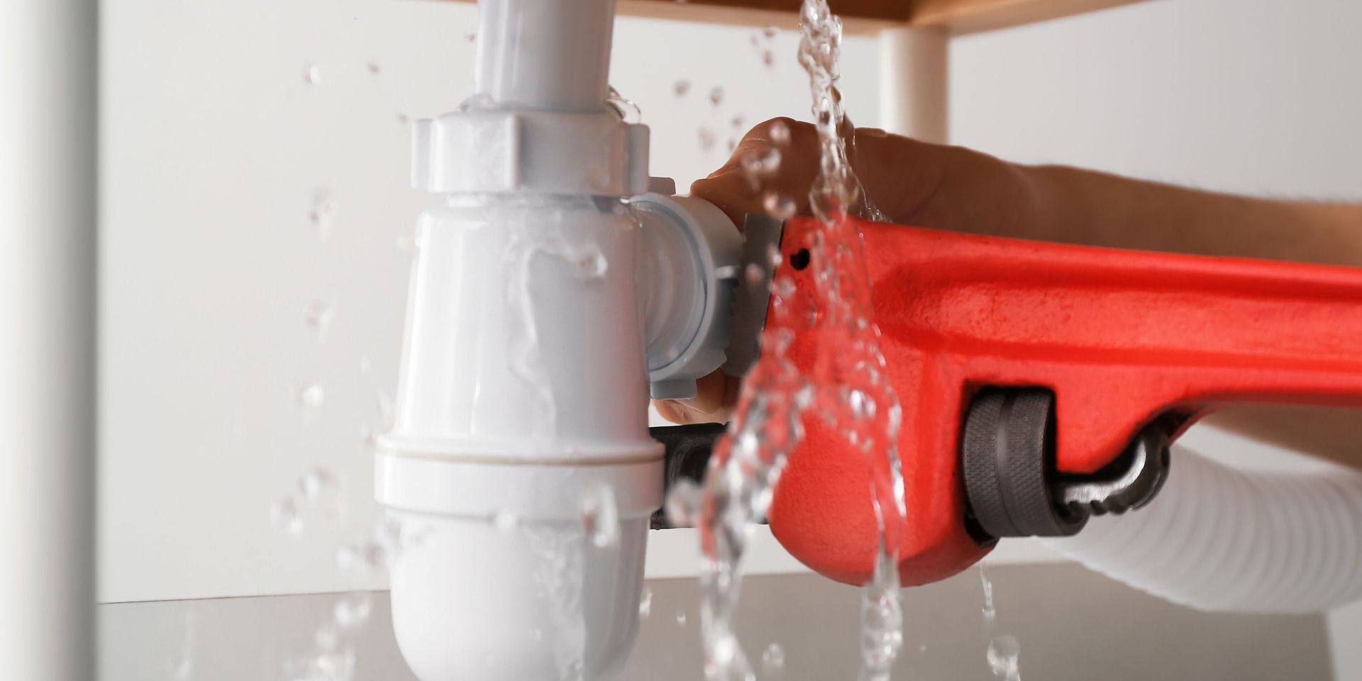A person using a red wrench on a leaking white pipe in a sink. Water streams out.
