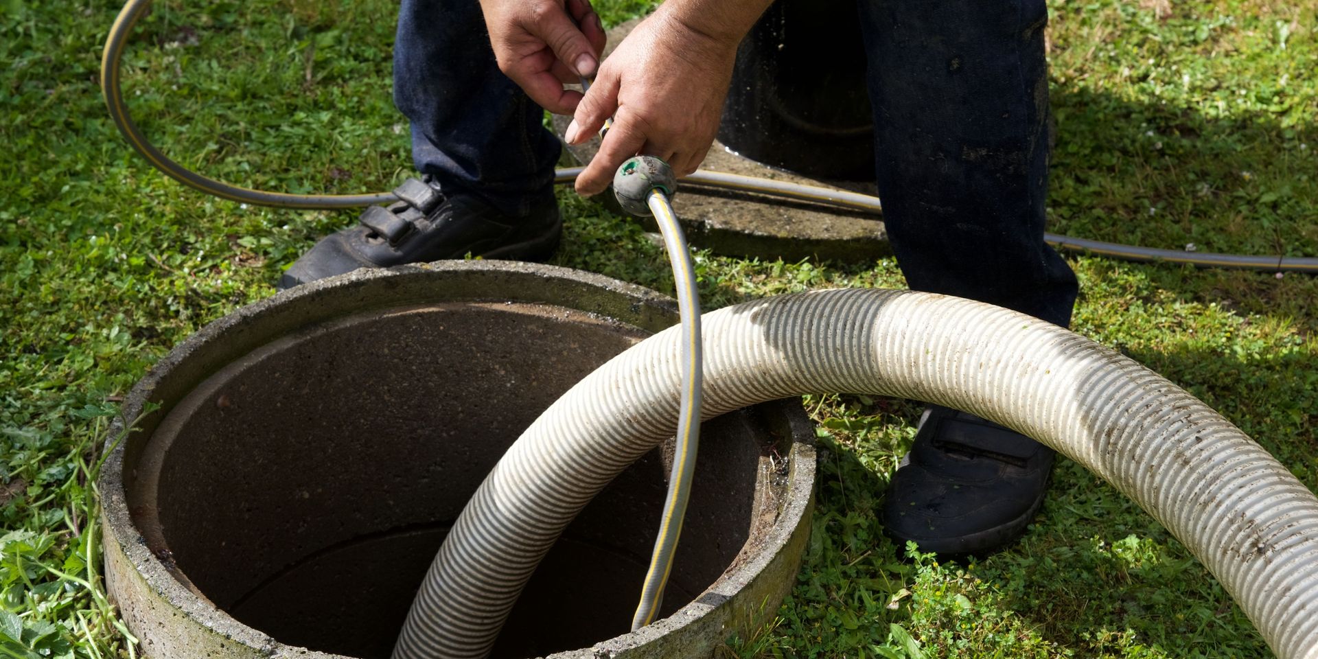 Person using a hose to service a septic tank; outdoors, green grass, concrete tank.