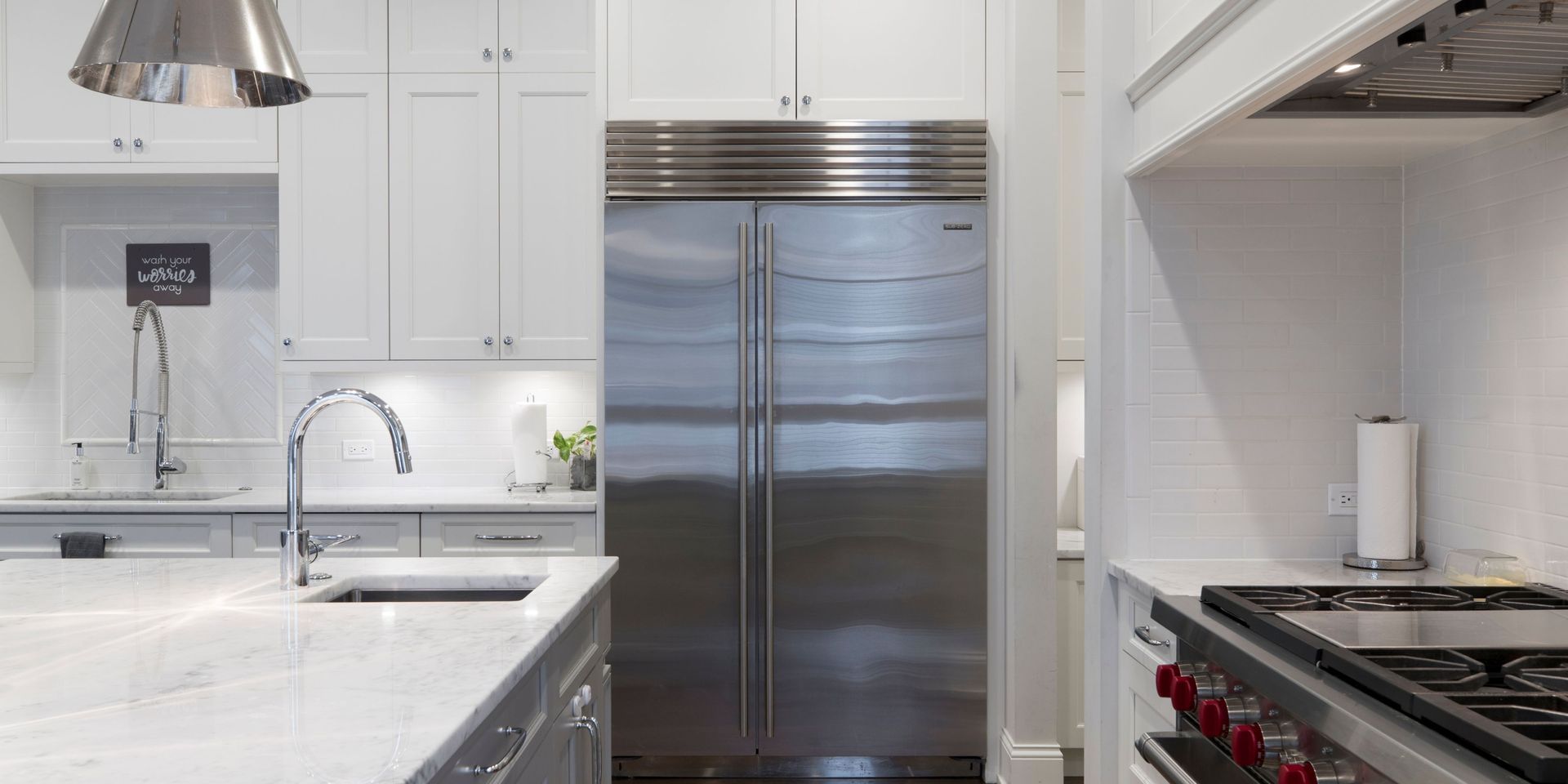 Stainless steel refrigerator centered in a white kitchen with marble countertops and a stovetop.