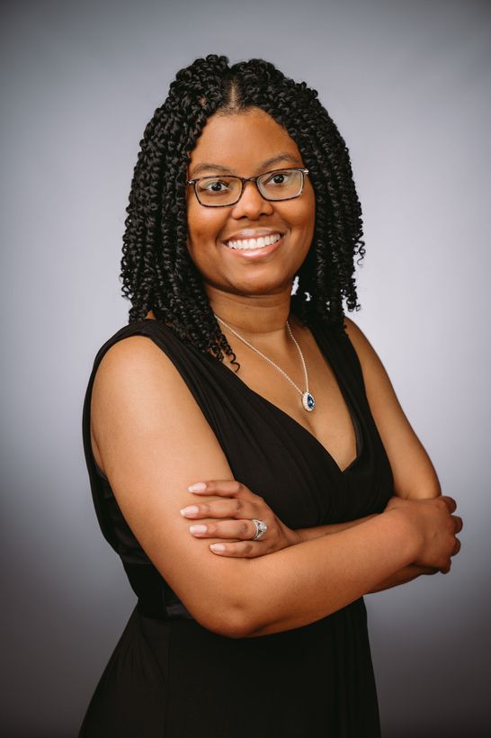 A woman wearing glasses and a black dress smiles with her arms crossed