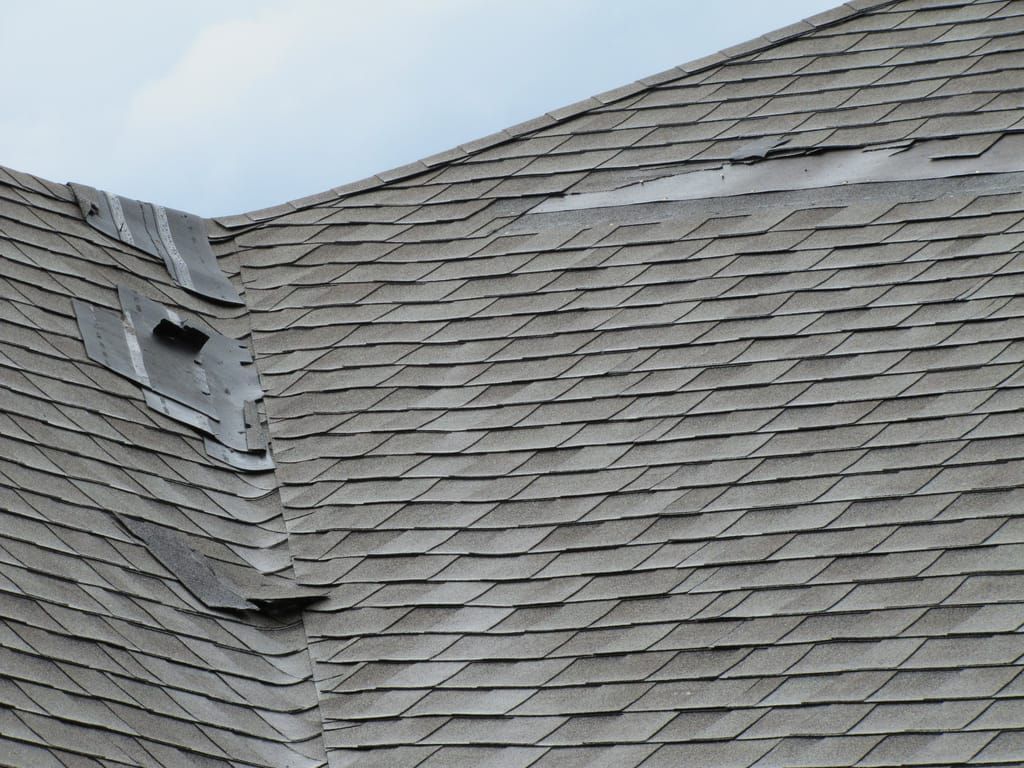 Damaged asphalt shingle roof with missing sections and exposed underlayment, overcast sky.