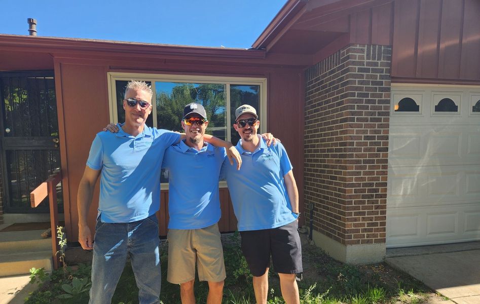 Three men in blue shirts pose in front of a house, one with arm around others, sunny day.