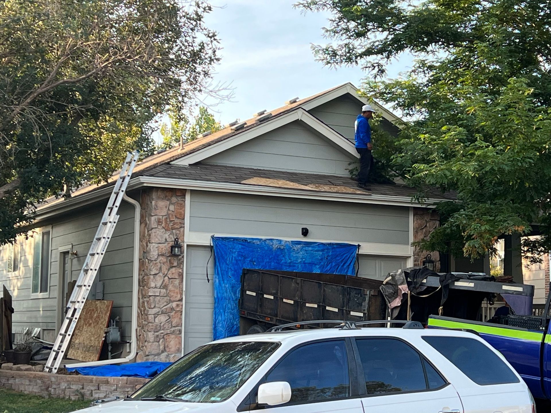 A person on a roof repairs shingles. A ladder leans against the house.