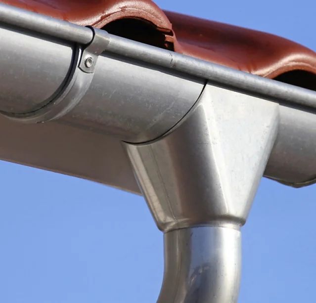 Silver gutter system with downspout, installed on a roof with brown tiles, against a blue sky.