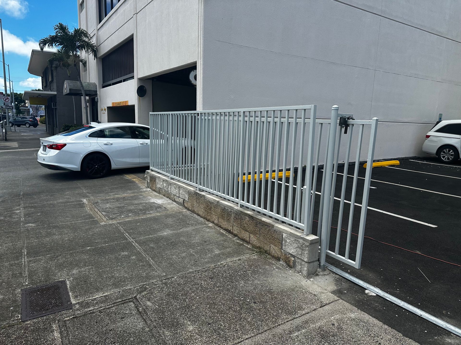 Metal gate and fence next to a building, partially blocking a parking area. A white car is parked nearby.