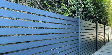 A modern, dark grey horizontal slat fence runs alongside a paved path with green trees in the background.
