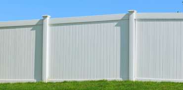 A white vinyl privacy fence stands against a clear blue sky above a green grass lawn.