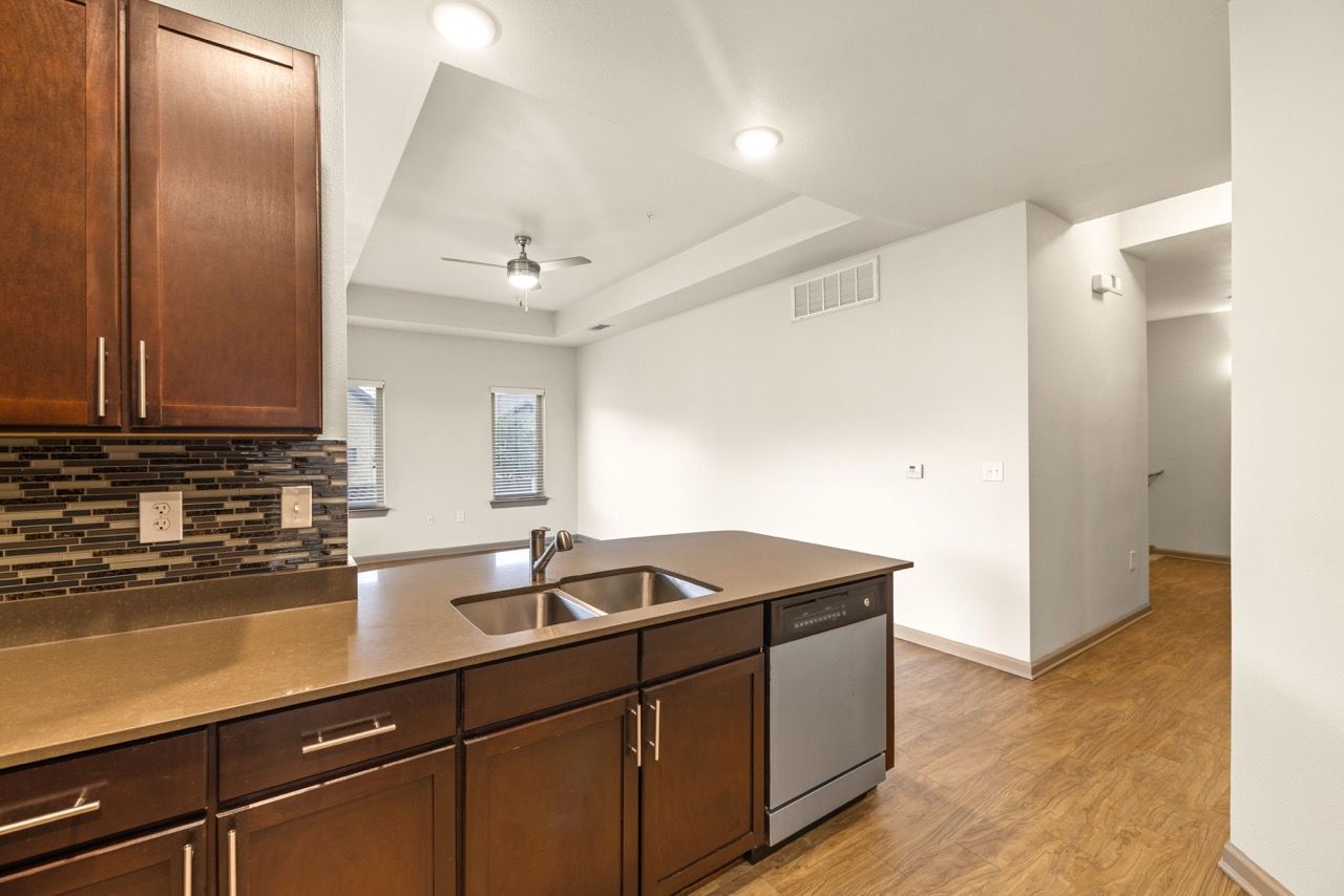 Open-concept kitchen with dark wood cabinets, a double sink island, stainless dishwasher, and mosaic tile backsplash.