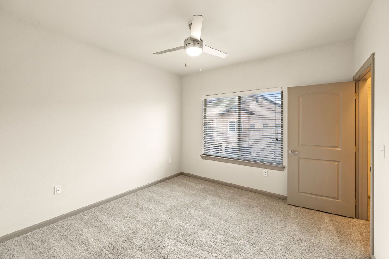 Bedroom interior in an apartment: beige walls, carpet, window with blinds, and a ceiling fan.