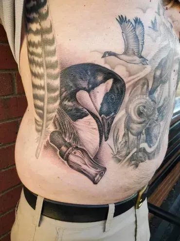A man has a tattoo of a duck and a duck call on his stomach.
