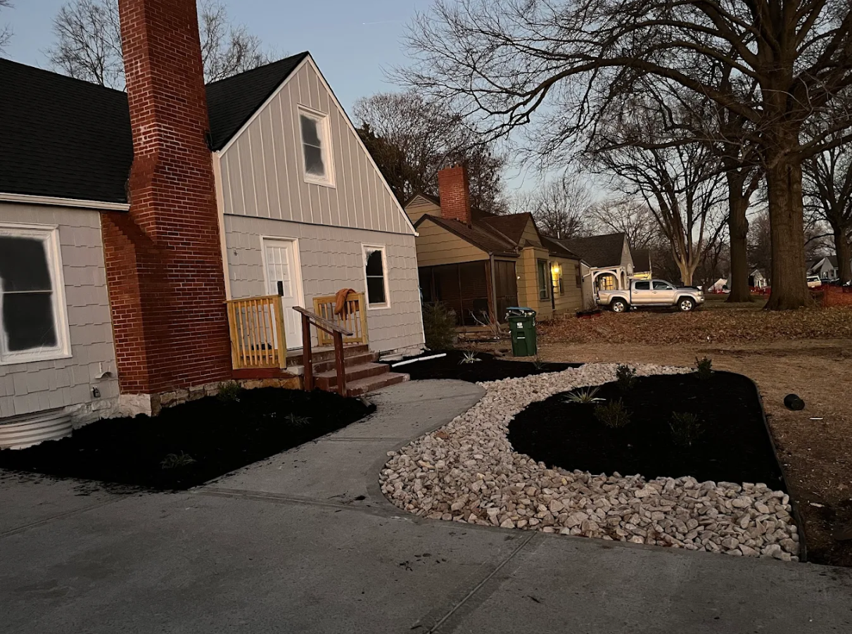 House with damaged asphalt driveway and stone landscaping beside a tree-lined yard