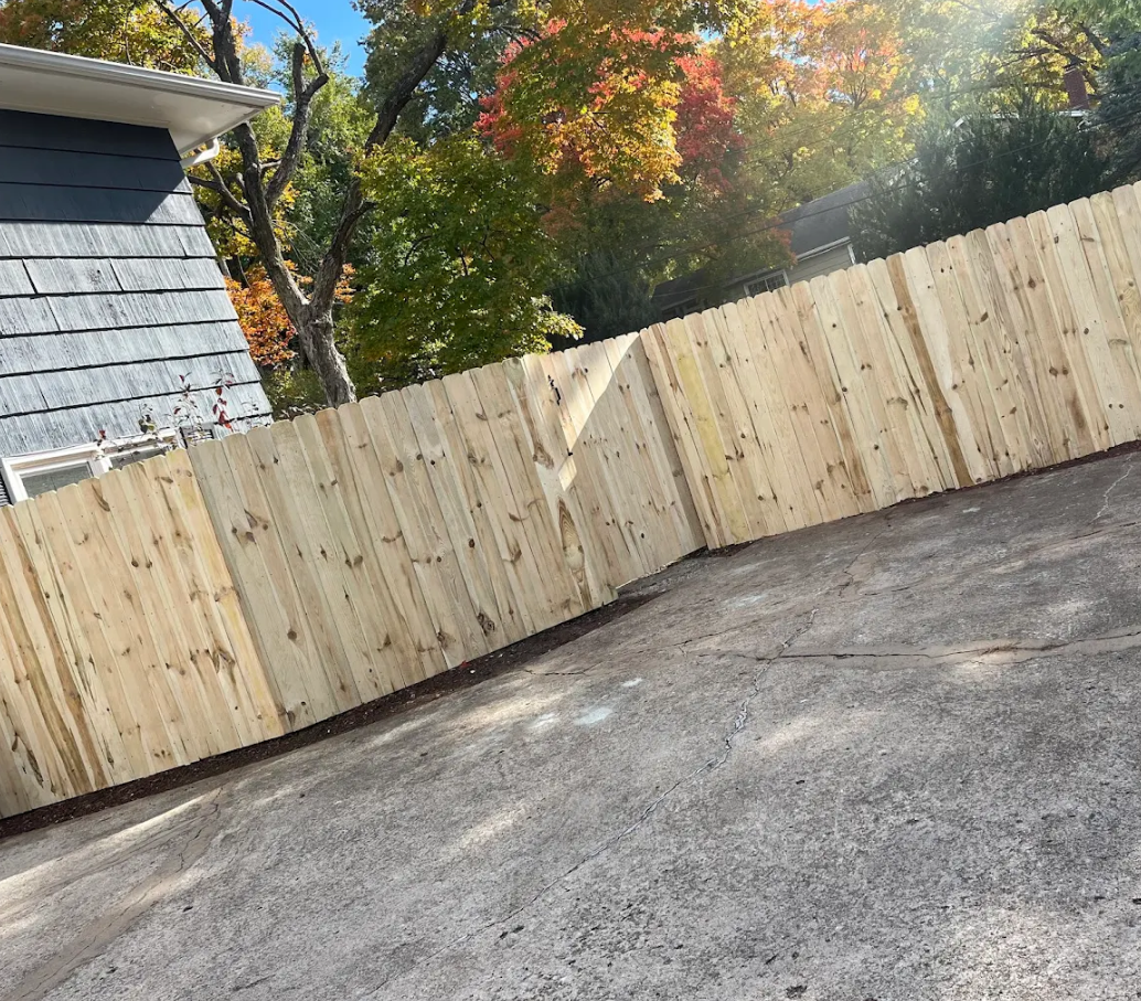 Empty paved driveway beside a light wooden fence and gray house, with autumn trees in the background
