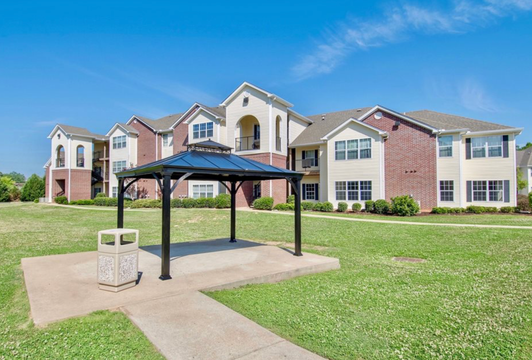 A gazebo is in the middle of a lush green field in front of a large apartment building.