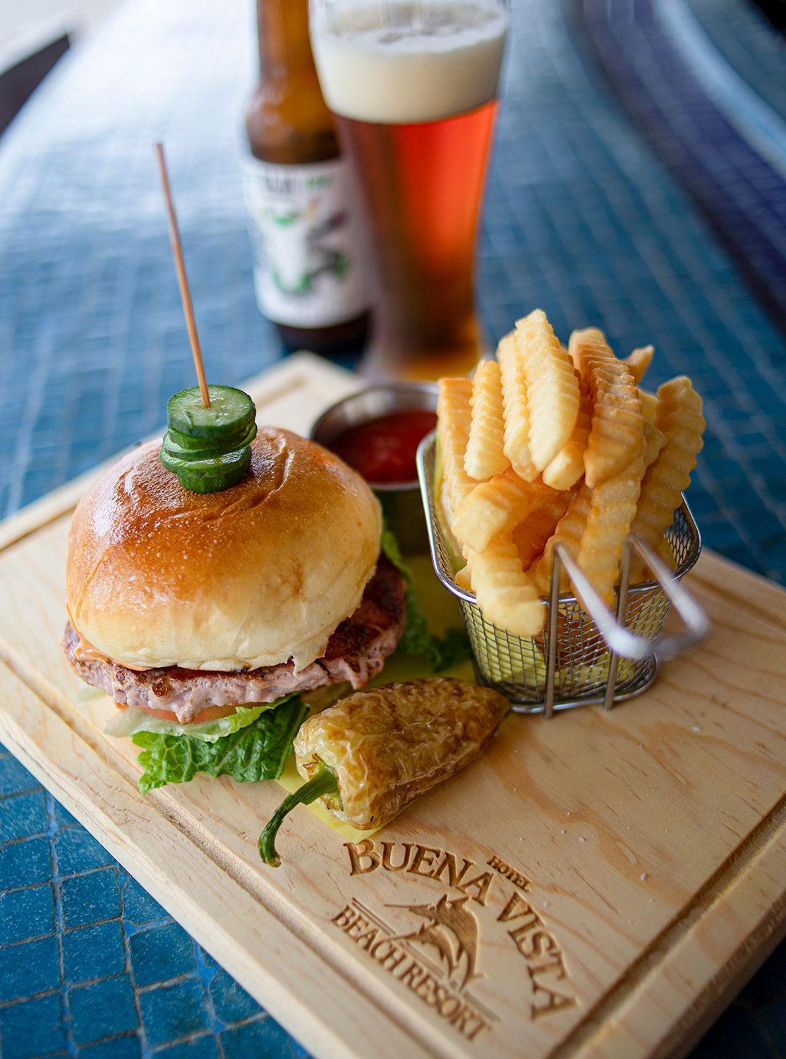Una hamburguesa y papas fritas sobre una tabla de cortar de madera con un vaso de cerveza al fondo.