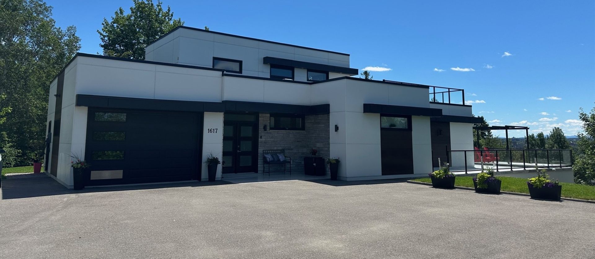 Une grande maison blanche avec une porte de garage noire est située au sommet d'une colline verdoyante.