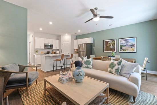 Open-concept living space with light green walls, white kitchen, beige sofa, rug, and wooden accents.