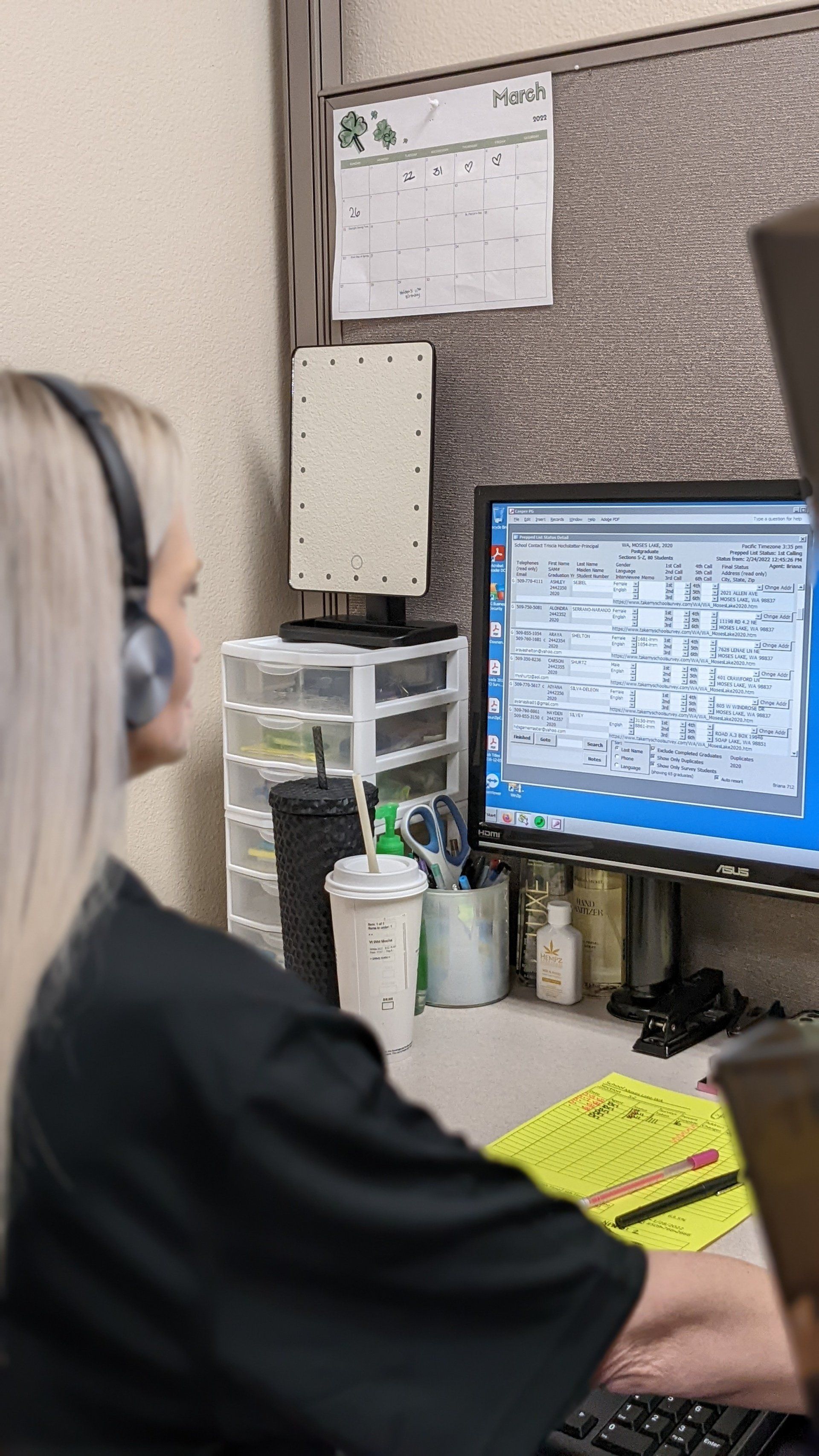 Woman with headphones at a desk, typing on a computer. A calendar and storage containers are visible.