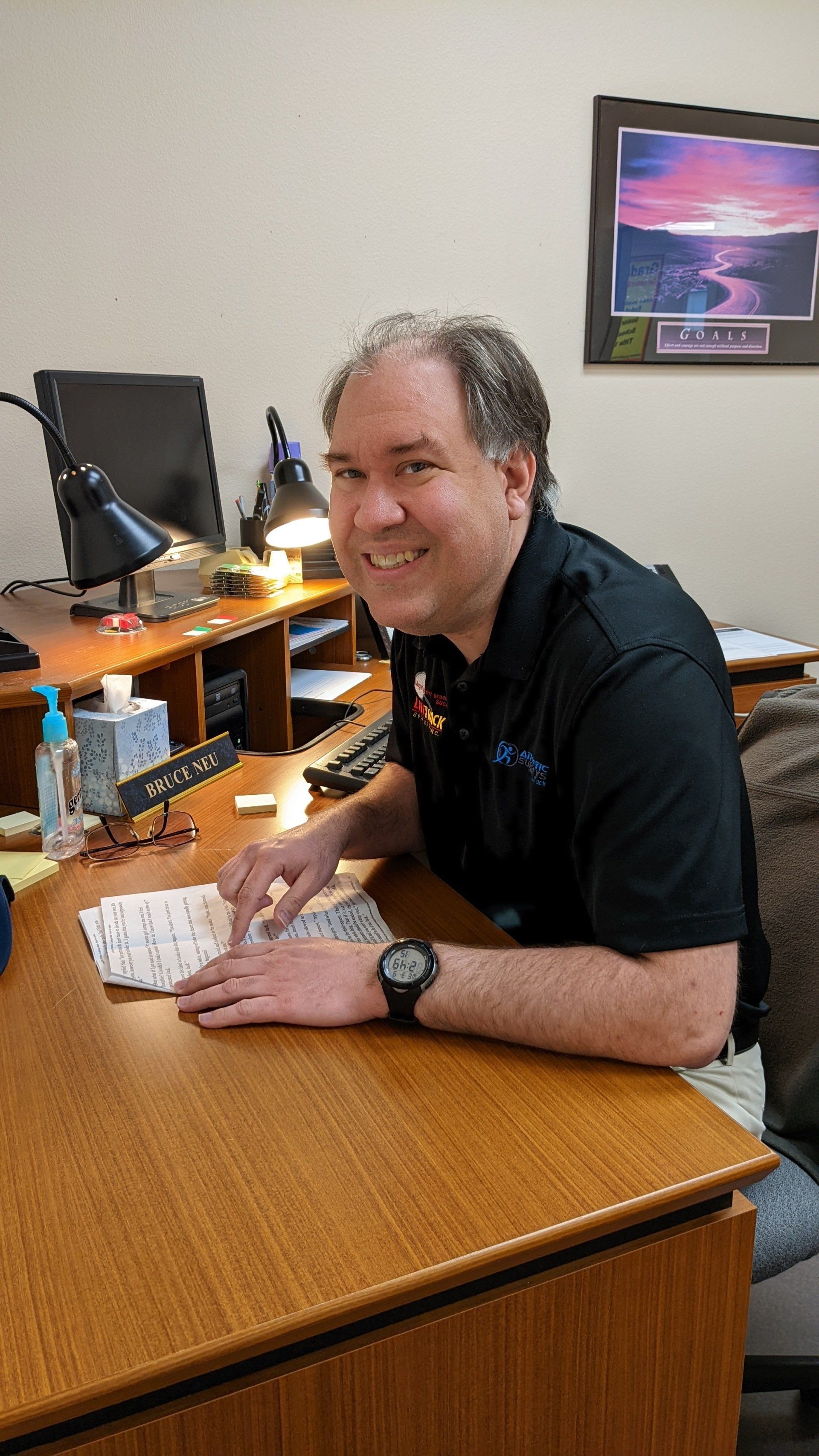 Man smiling at desk, wearing black shirt. Desk has monitor, lamp, phone, hand sanitizer, and art.