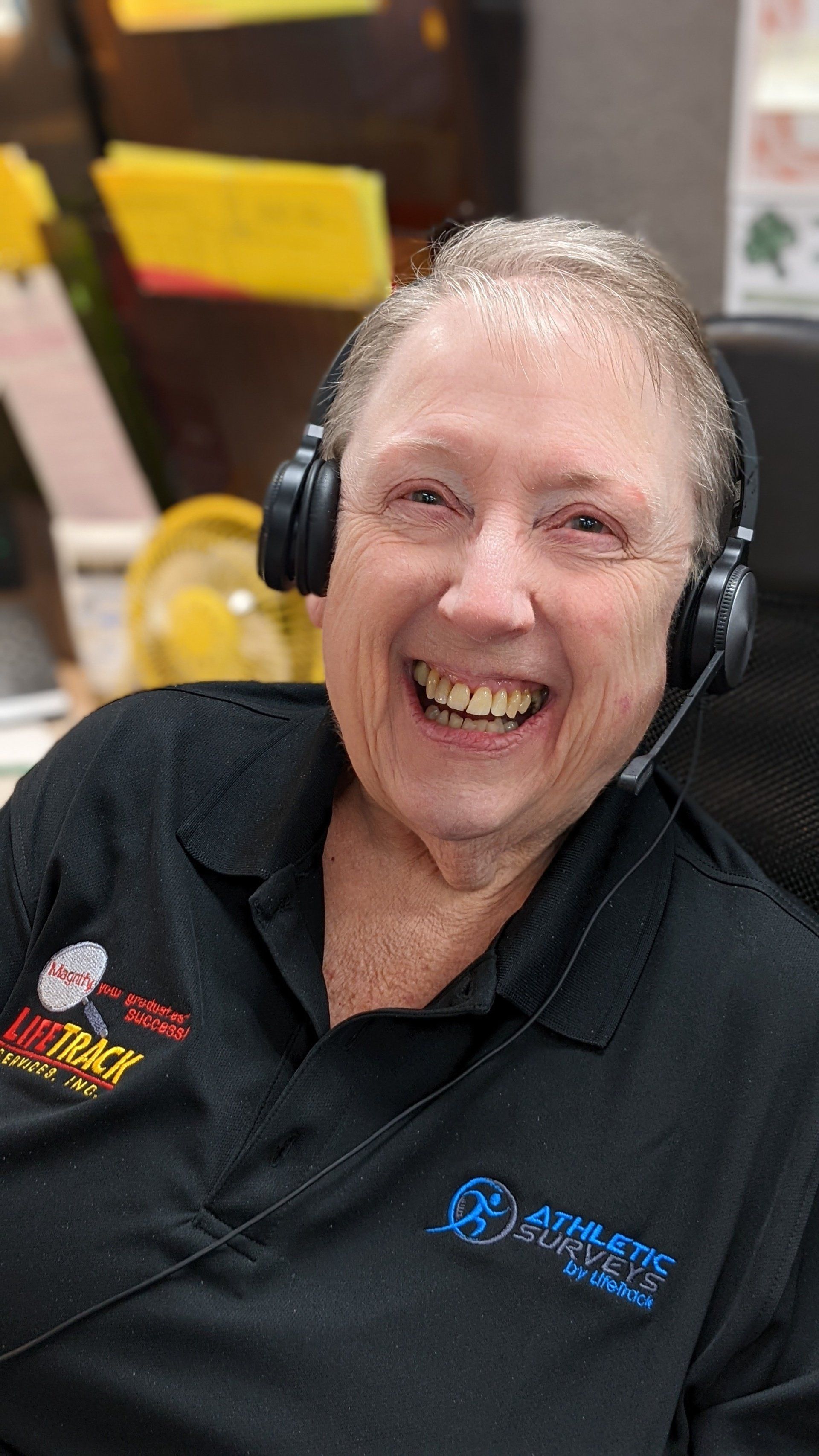Woman wearing a headset, smiling widely in an office setting. Wearing a black shirt with logos.