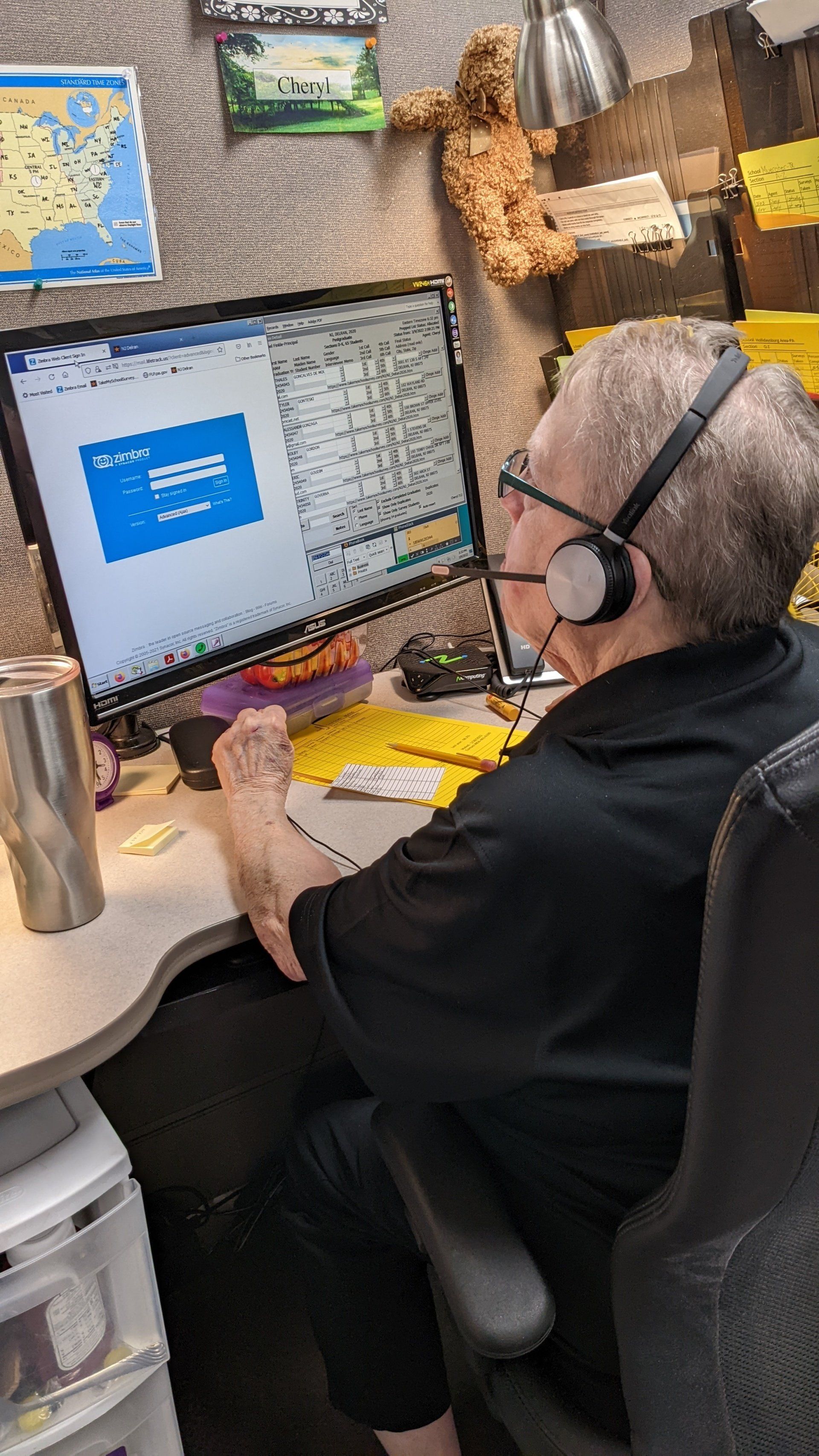 Person wearing headset at computer, appearing to work. Gray hair, black shirt, desk setup with drink and papers.