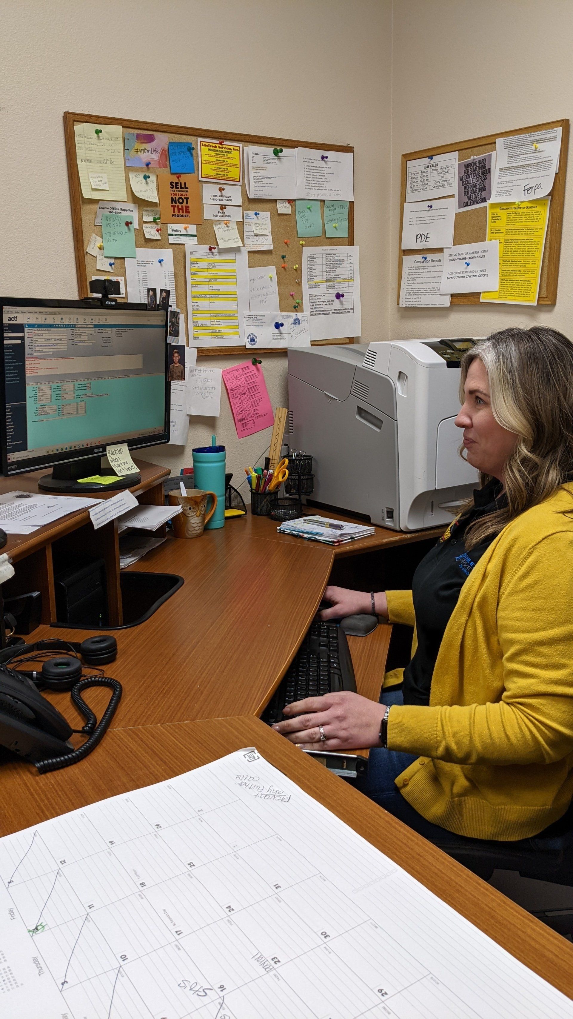 Woman at desk, typing on computer, with bulletin boards and printer visible.