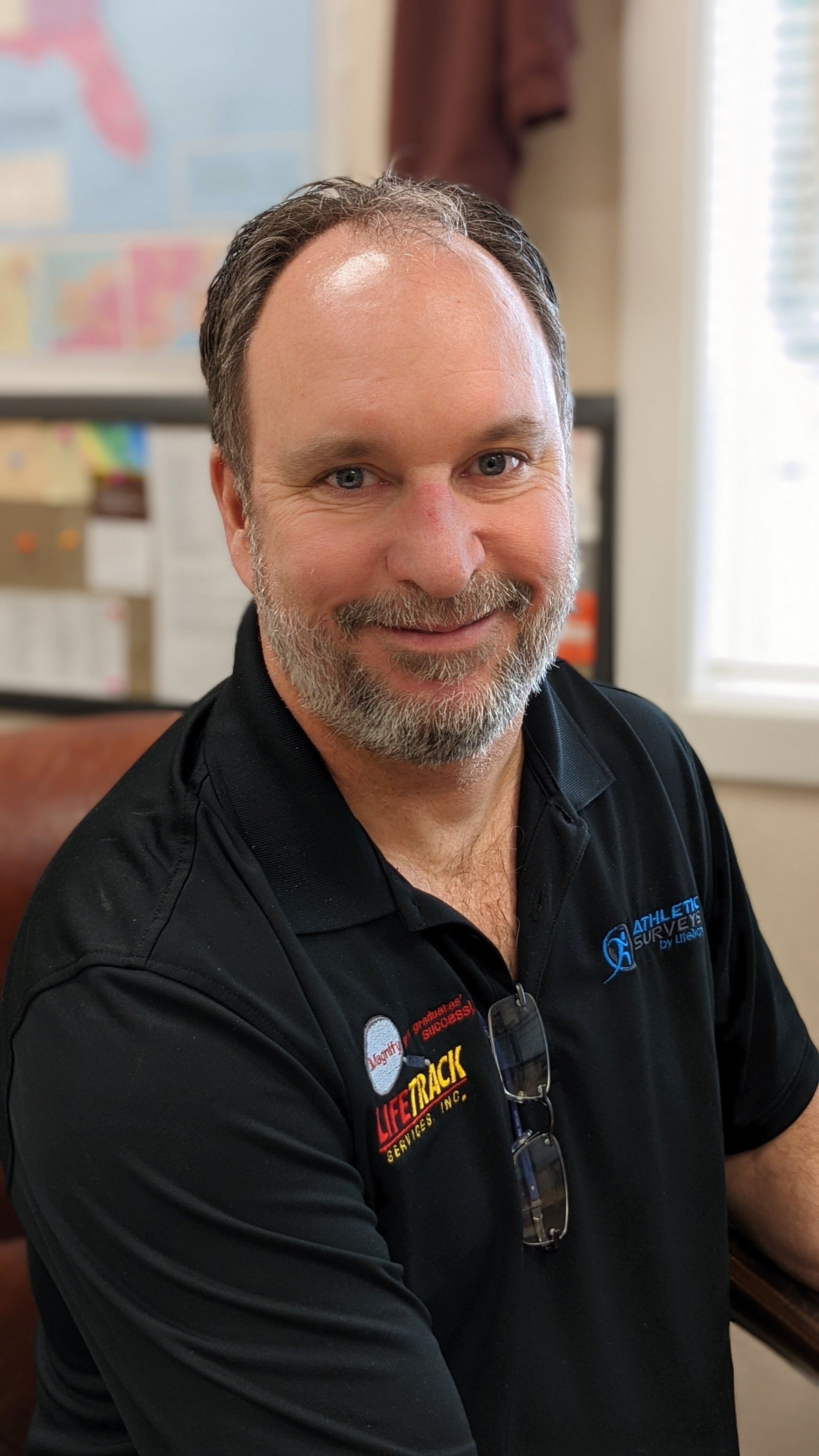 Man with a graying beard smiles, wearing a black shirt. Office setting with framed map and window.