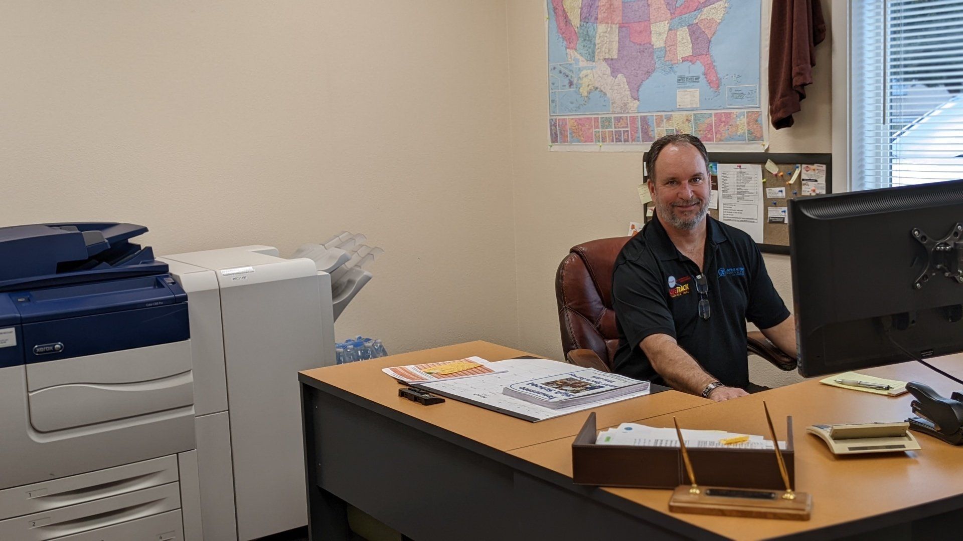 Man sitting at a desk in an office with a printer, monitor, and map on the wall.
