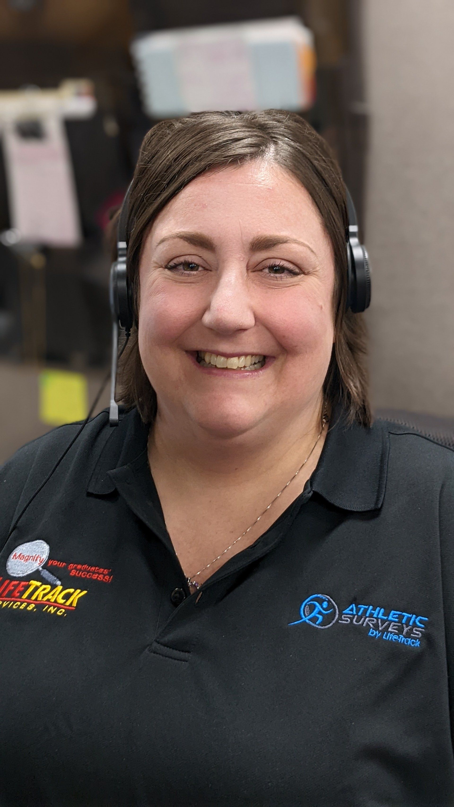 Woman wearing a headset, smiling, in a black shirt with company logos, indoors.