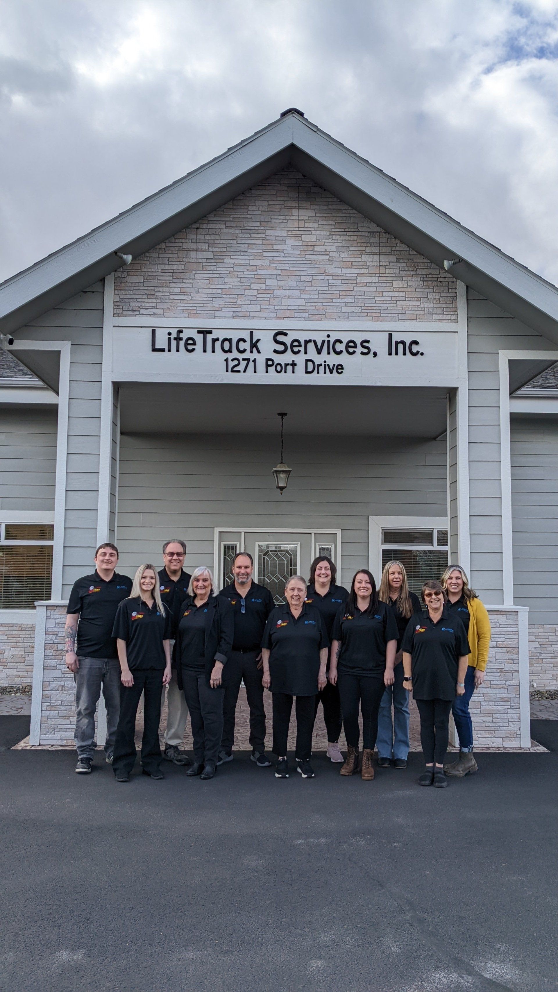 Life Track Services, Inc. staff pose in front of their office building. Grey building, cloudy sky, people in black shirts.