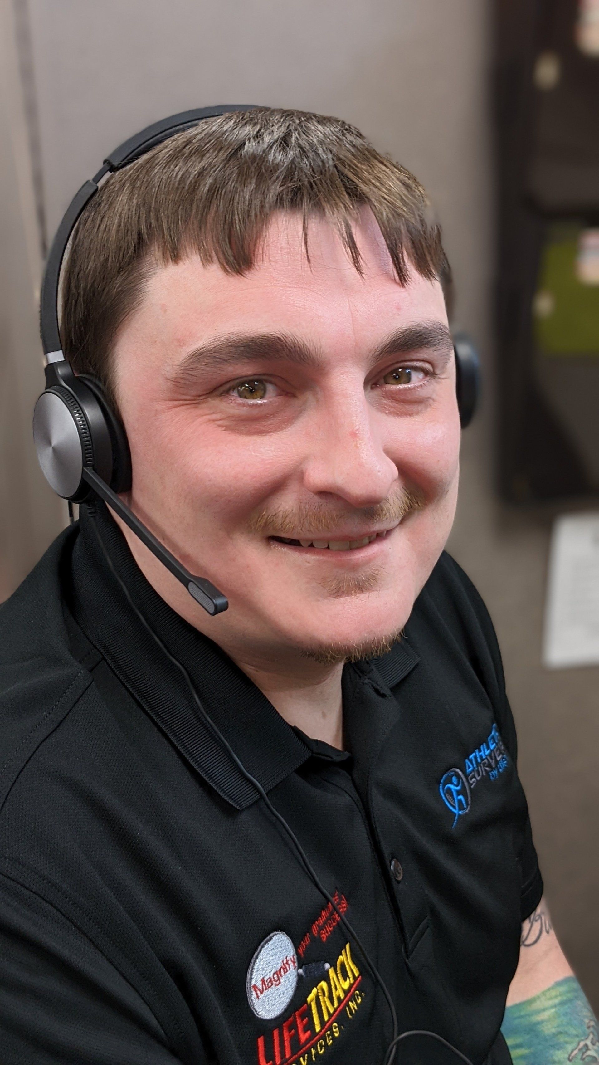 Man wearing headset, smiling at the camera. He wears a black polo shirt, possibly in an office setting.