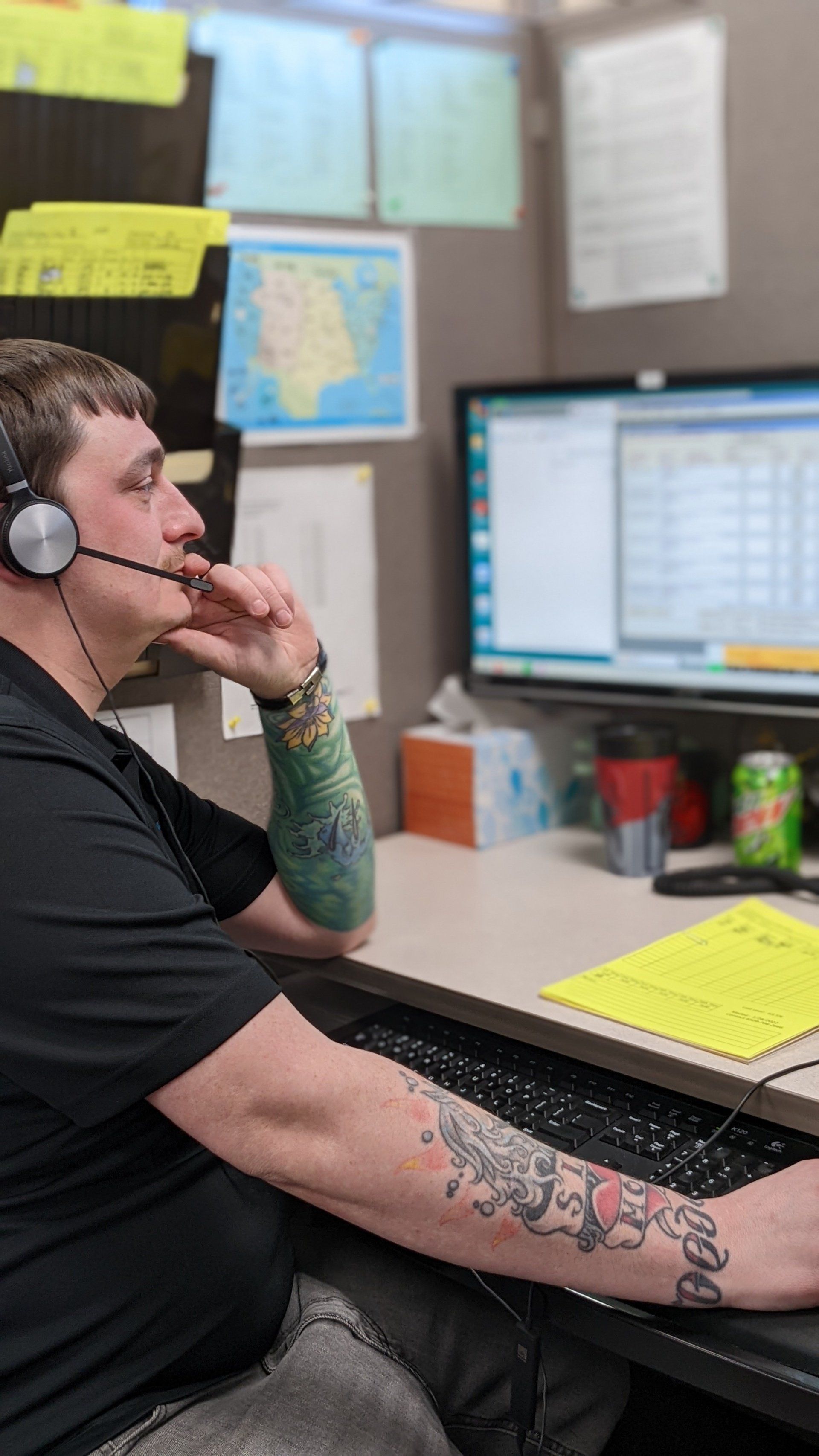 Man with headset at a computer in a cubicle, looking at the screen, with a tattooed arm.
