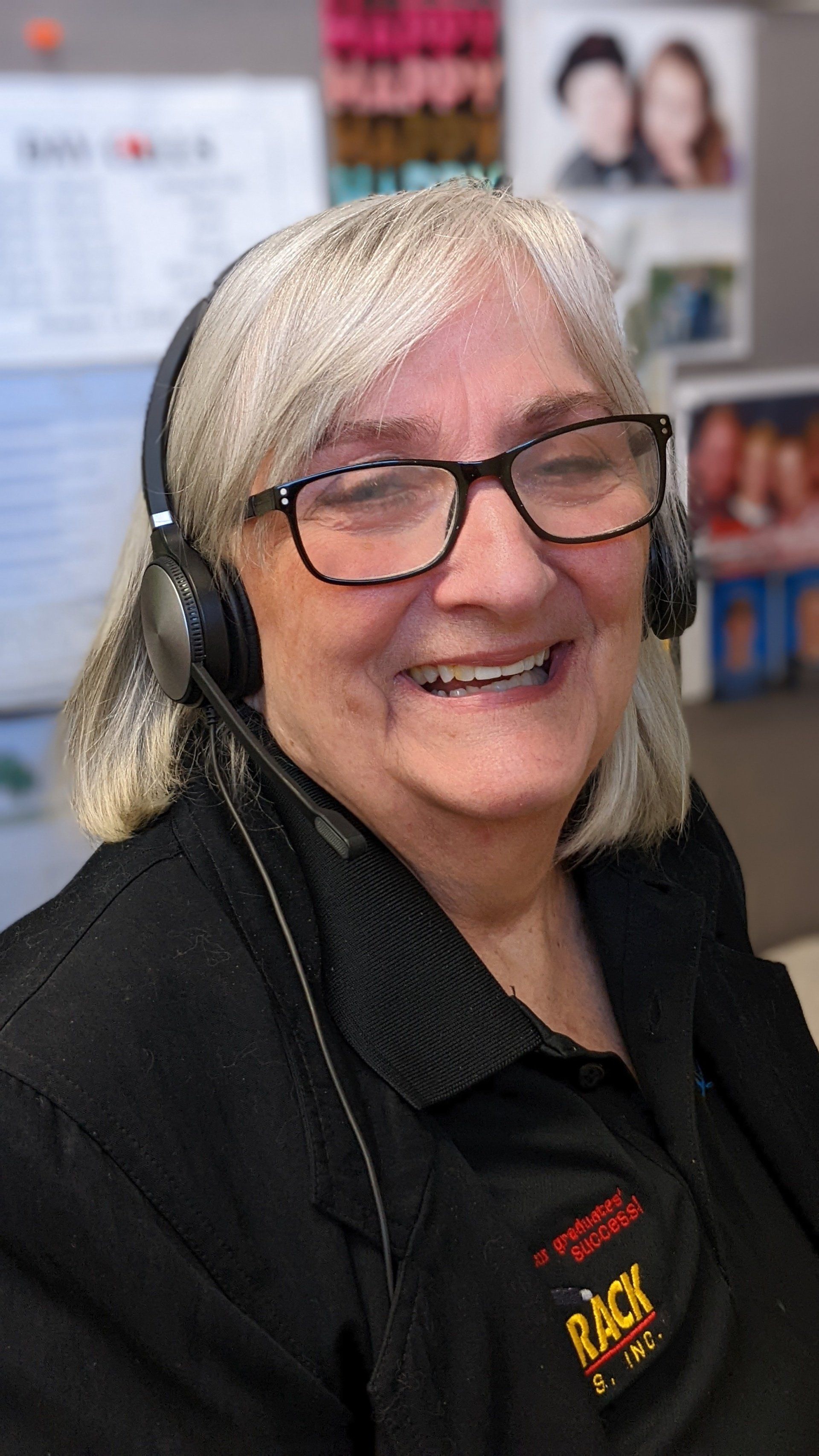 Woman with glasses and headset, smiling at desk. Black shirt with logo visible.