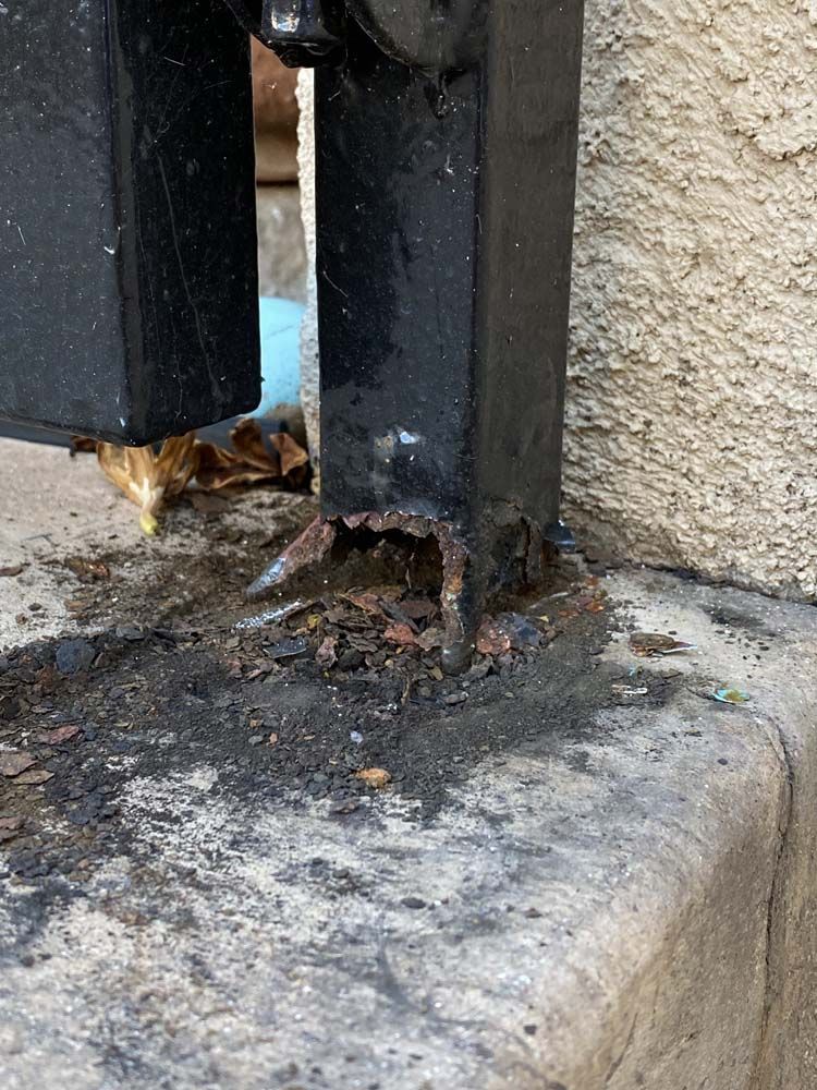 Corroded black metal gate post on concrete, showing rust damage at the base.