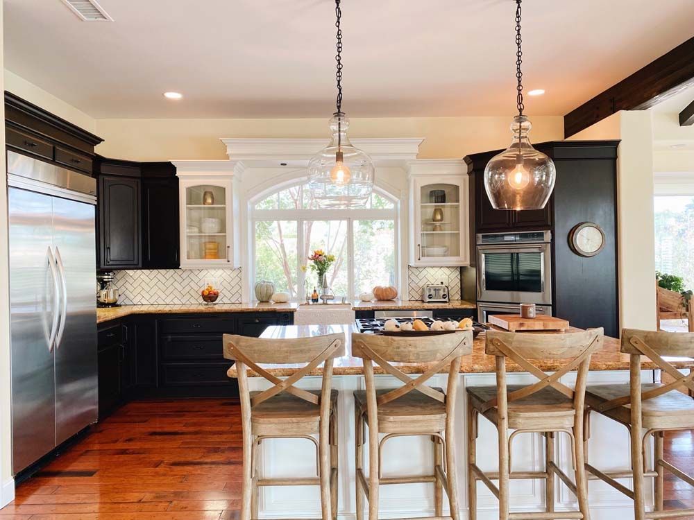 Kitchen with black and white cabinets, island with stools, pendant lights, stainless steel appliances, and wooden floors.