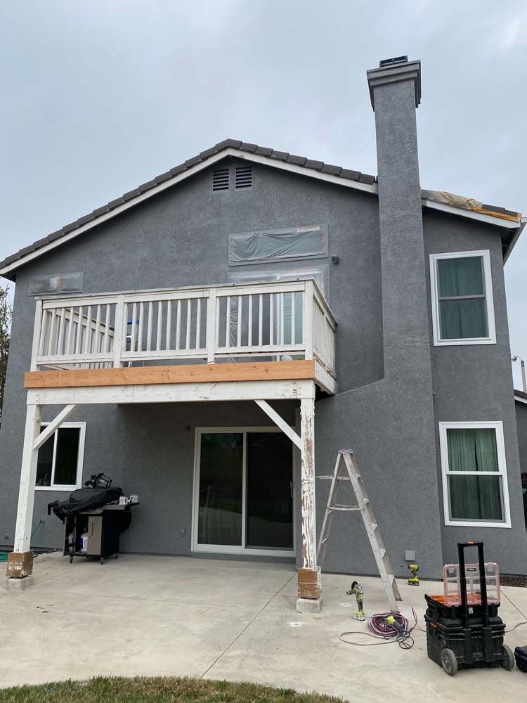 Back of two-story house with gray stucco, white deck, and chimney. Construction tools present. Overcast sky.