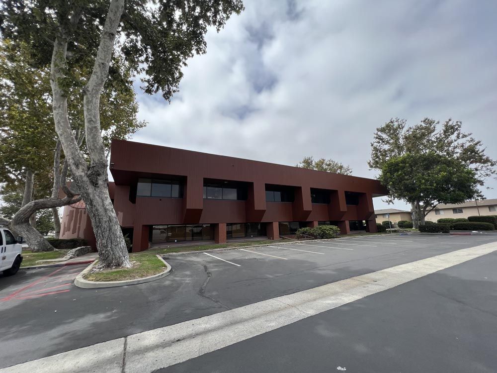 Brown two-story office building with large windows. Trees frame the building. Cloudy sky.