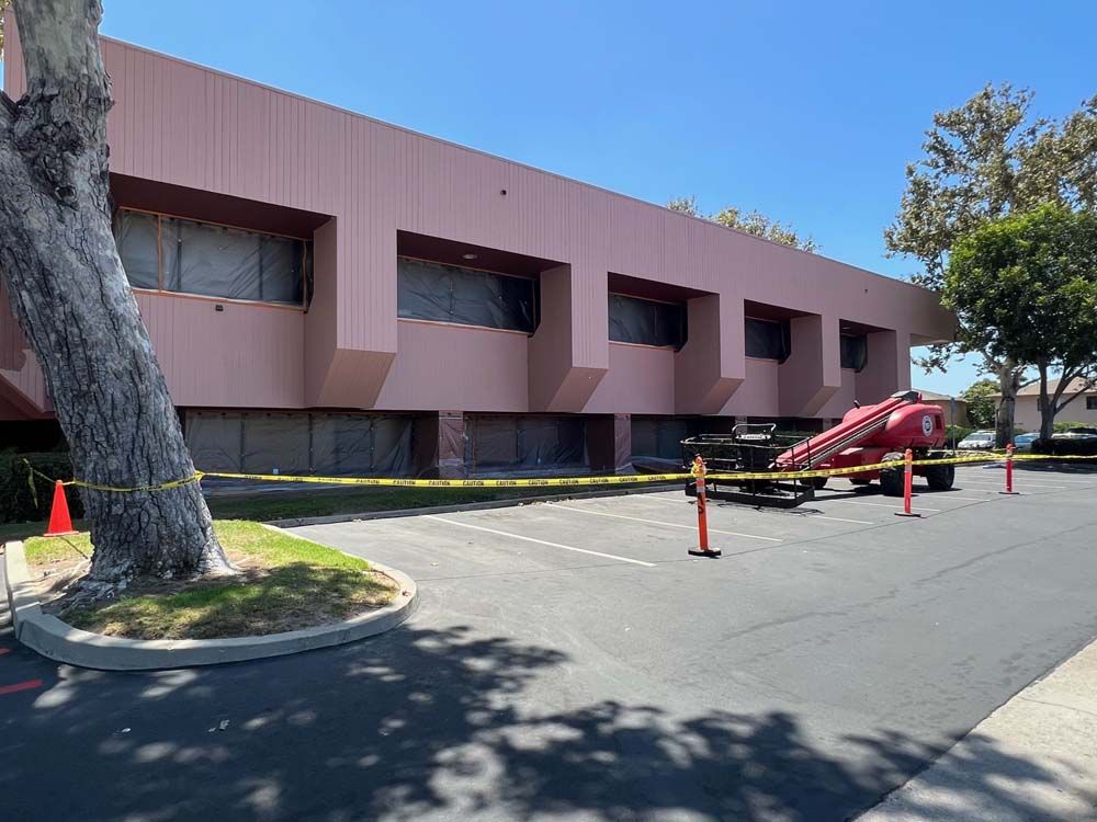 Pink building with covered windows, construction equipment, and caution tape in a parking lot.