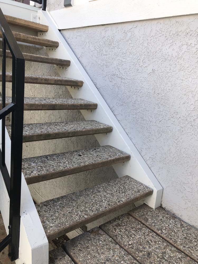 Exterior staircase with gray stone-like treads and white risers, black railing, beside a stucco wall.