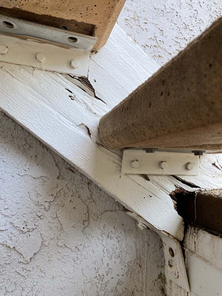White painted wooden beams with rust colored brackets connected to a brick structure; wood damage is visible.