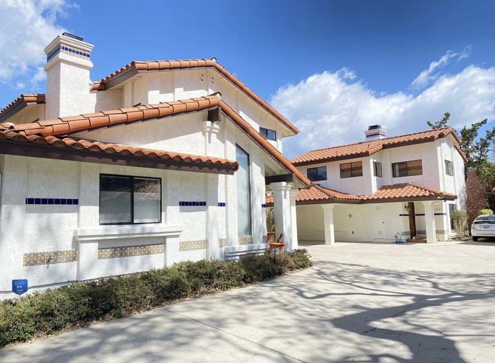 White stucco house with red tile roof, blue accent tiles, and a driveway on a sunny day.