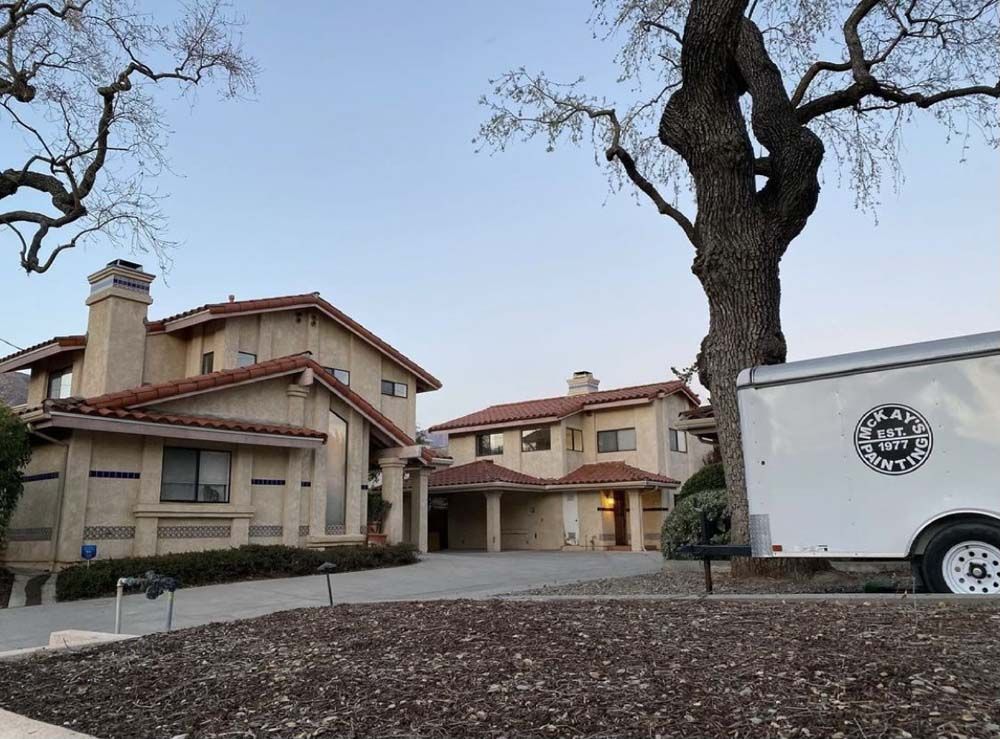 Two-story beige homes with red tile roofs. A white trailer is parked beside a large tree.