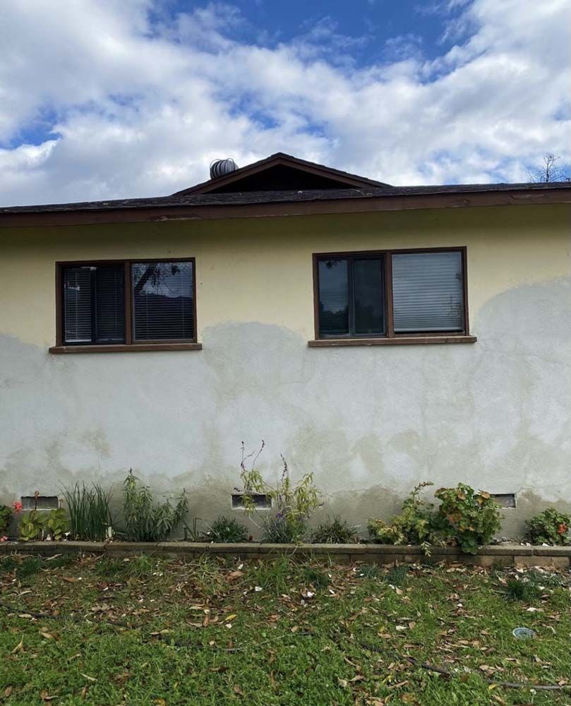 Exterior of a home with a light yellow wall, two windows, and a brown roof against a cloudy sky.