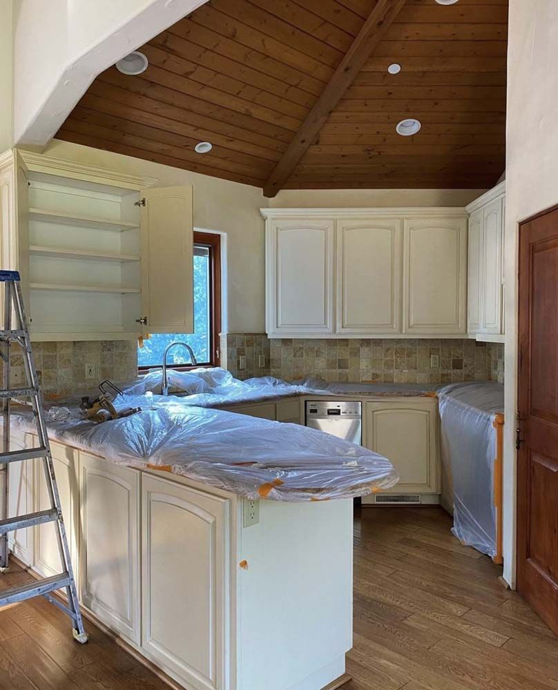 Kitchen renovation in progress: white cabinets, wooden ceiling, countertop covered in plastic, ladder, and wood flooring.