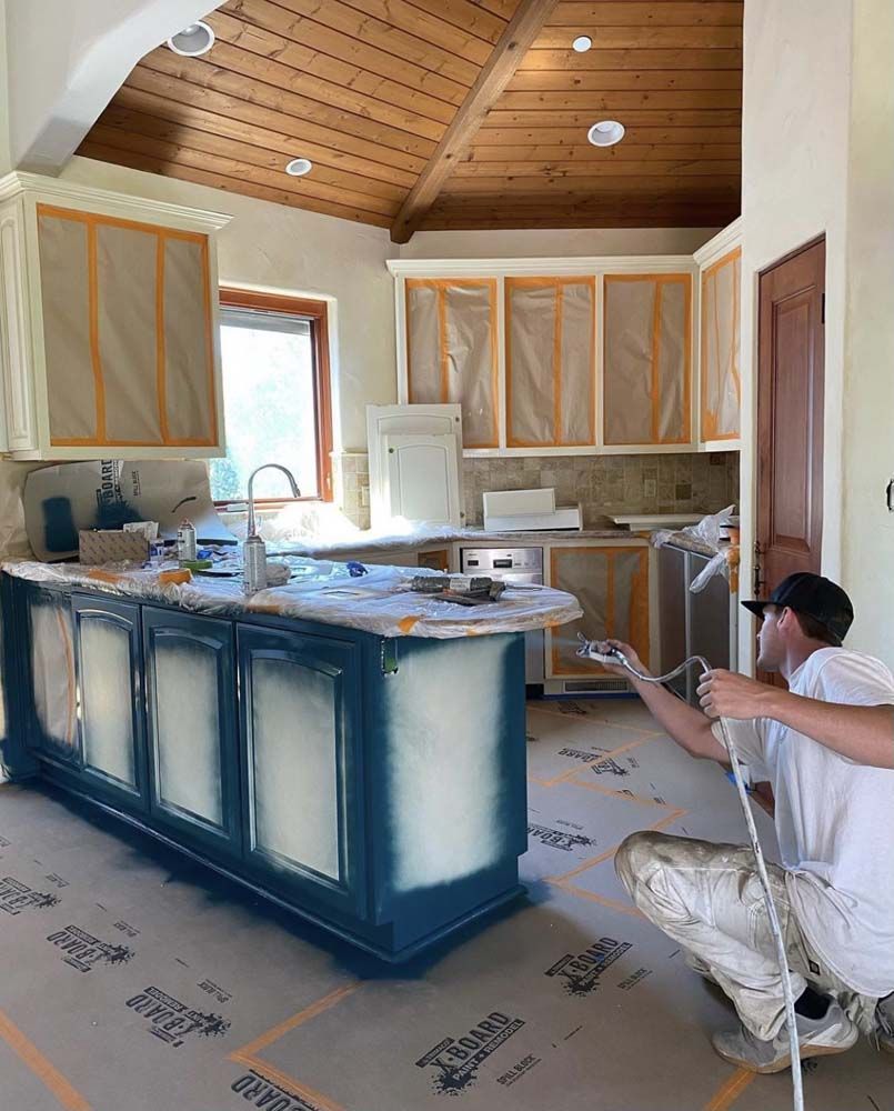 Person painting blue kitchen cabinets with a sprayer. Room has light-colored walls, wood ceiling and countertops.