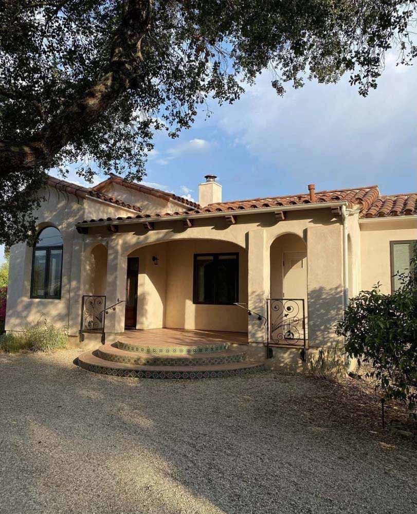 Spanish-style house with arched porch and tiled roof, set in a gravel driveway with tree shade.