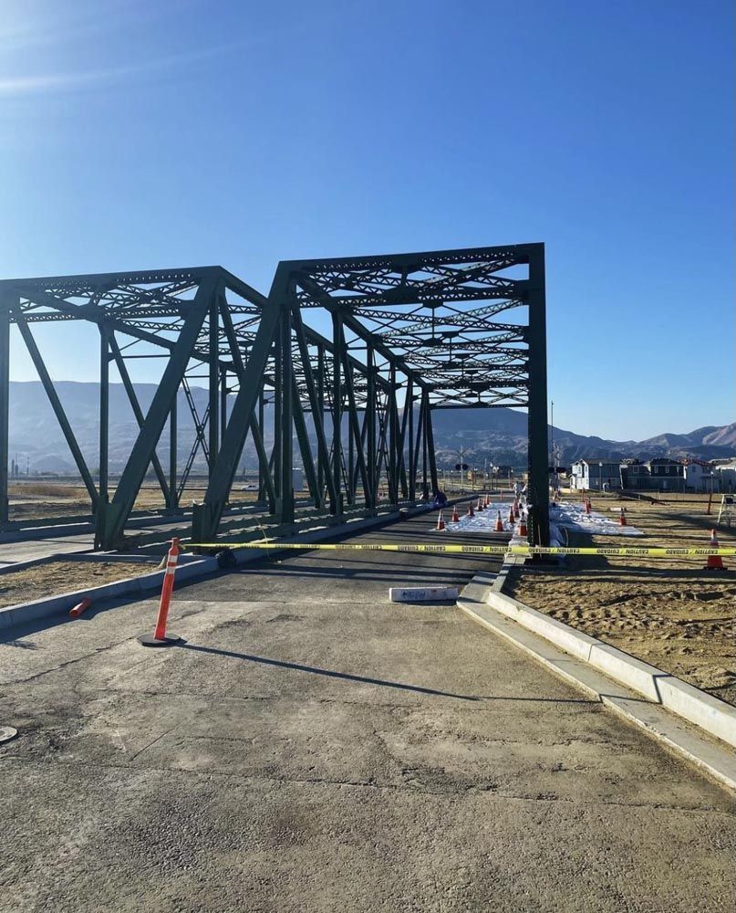 Green steel truss bridge over a paved area, with mountains in the background under a clear blue sky.
