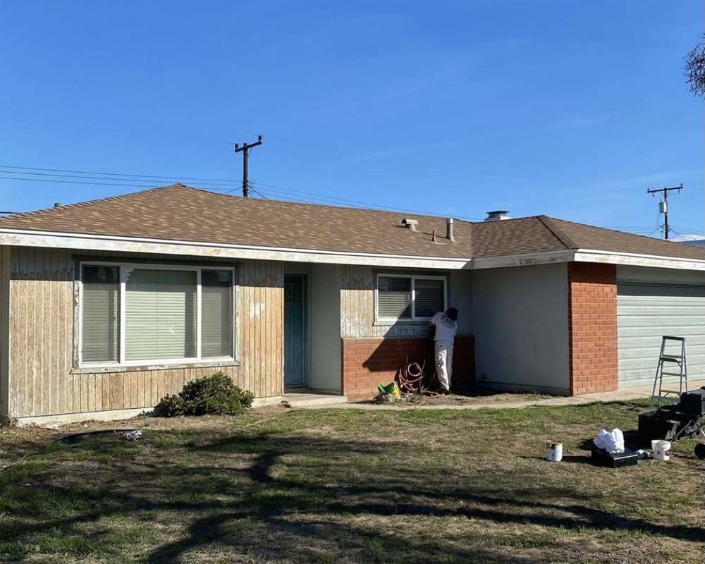 House being painted; person in white painting window. Brown roof, blue sky, and green grass.