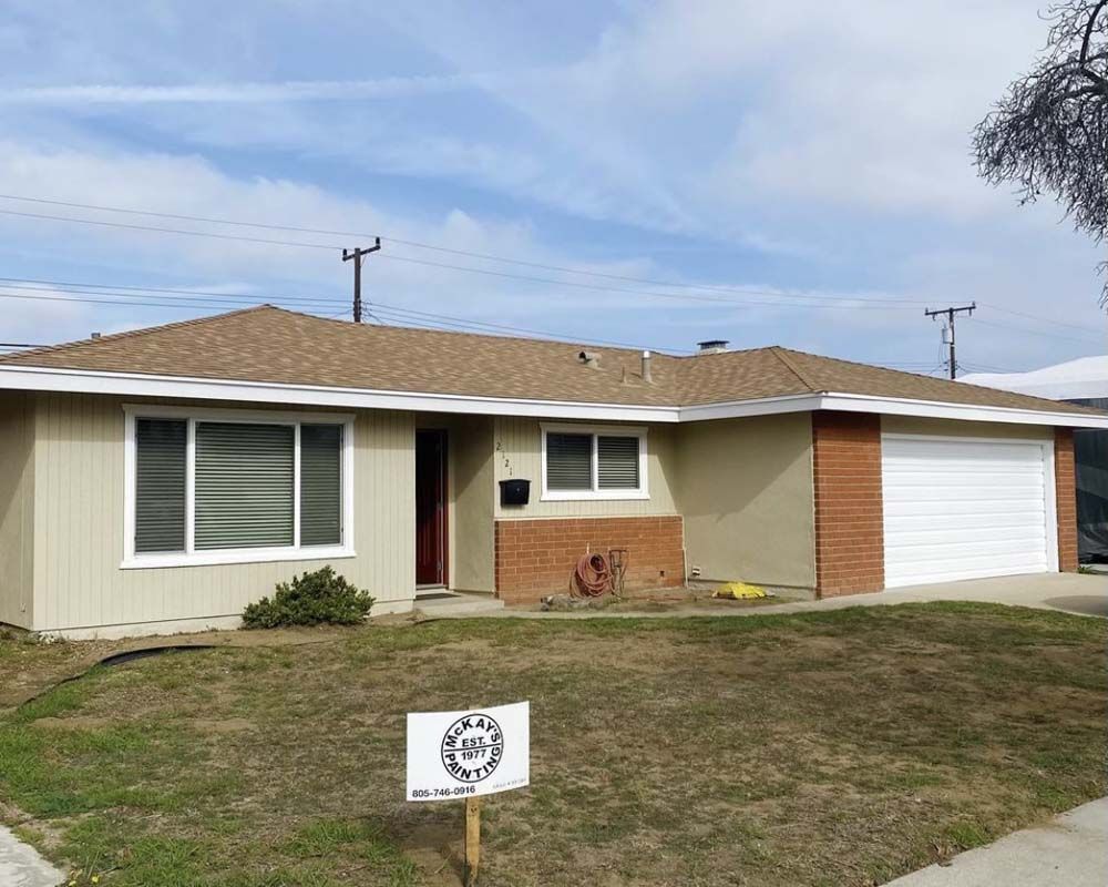 Beige ranch-style house with brown roof, brick accents, white garage door, and a sign in the front yard.